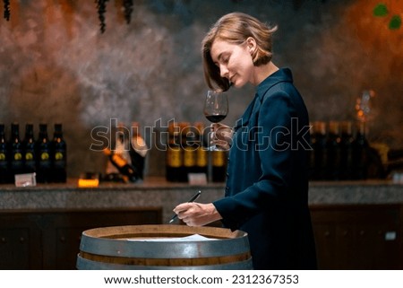Professional woman sommelier testing red wine in wine glass with tasting and smelling at wine cellar with wooden barrel in wine factory. Winery liquor manufacturing industry and winemaker concept. Royalty-Free Stock Photo #2312367353