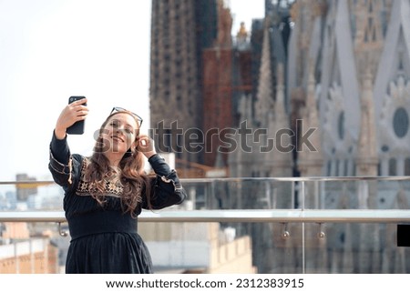 Happy young Latin woman, tourist in traditional Mexican dress with sunglasses smiling and taking selfie on smartphone against historic the building Sagrada Familia in Barcelona during sightseeing trip