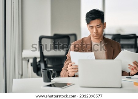 Serious busy young Asian professional business man executive ceo manager sitting at desk in office working checking corporate financial accounting documents feeling worried about taxes or bills. Royalty-Free Stock Photo #2312398577