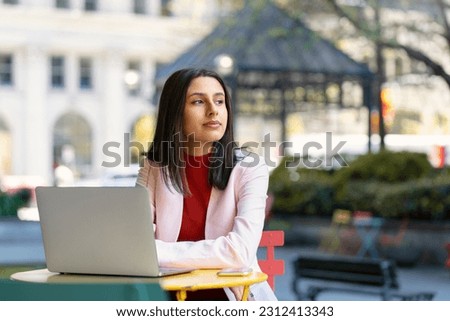 Portrait of beautiful pensive Indian woman using laptop computer, planning project looking away waiting someone on the street. Asian student studying in university campus using wireless internet 
