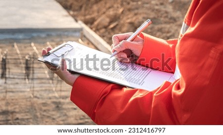 Action of a civil engineer is checking the building quality report form with construction work site as background. Industrial working action scene with people, close-up and selective focus.