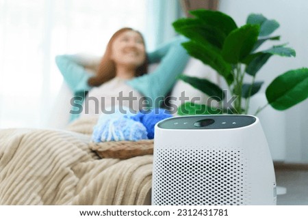 Happy Asian young woman turning on high efficiency air purifier while staying and relaxing in the living room. Woman using air purifier in her house. Royalty-Free Stock Photo #2312431781