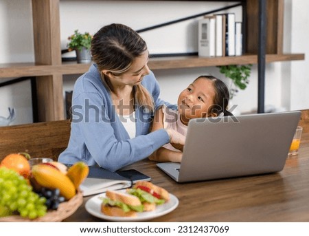 Asian young female mother and little cute girl daughter sitting smiling learning studying doing homework online via laptop notebook computer drinking orange juice eating sandwich together at home.