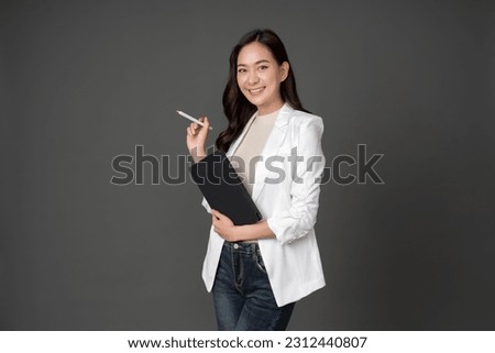 Asian female executive with long hair Smiling and giving outstanding eyes Working with a tablet and a pen wearing a white suit and stand to take pictures with a gray scene in the studio Royalty-Free Stock Photo #2312440807