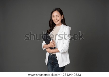 Asian female executive with long hair standing hugging a tablet for work, holding a pen, with a beautiful smile. wearing a white suit and stand to take pictures with a gray scene in the studio Royalty-Free Stock Photo #2312440823