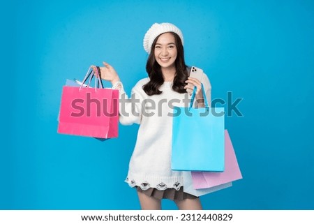 Asian woman shopaholic with long hair Take pictures in the blue studio scene Hold a shopping paper bag in your left and right hand. have a mobile phone wearing a white coat wearing a hat