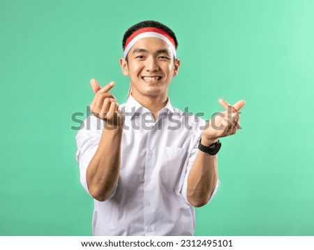 A portrait of an Asian man wearing a white shirt and a red and white headband, forming a Korean heart shape with his hands in a pose, isolated on a green background.