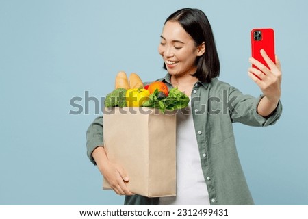Young fun woman wear casual clothes hold brown paper bag with food products do selfie shot mobile cell phone isolated on plain blue background studio portrait Delivery service from shop or restaurant