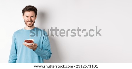 Celebration. Handsome young man celebrating birthday, holding bday cake with lit candle and smiling, making wish, standing over white background.