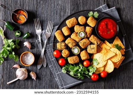 crispy fried jalapeno poppers with melted cream cheese filling with taco chips, tomatoes and salsa on black platter on dark wood table, horizontal view from above, flat lay