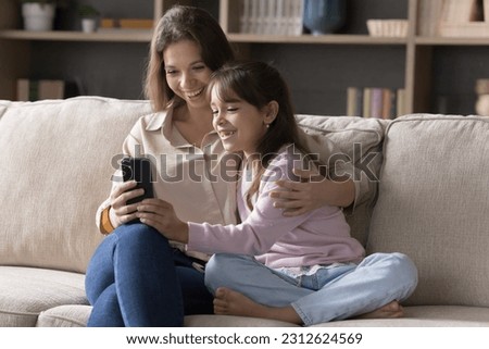 Cheerful mommy and pretty little kid girl using online application on mobile phone for Internet communication, video call, taking selfie picture, smiling, laughing, sitting close on sofa