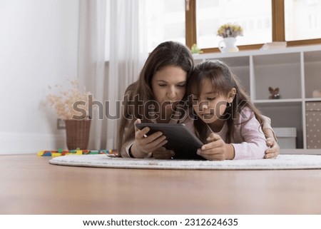 Focused young mom and serious little kid girl resting close on heating floor, using educational app on tablet, playing online learning game, reading book on screen, enjoying family leisure
