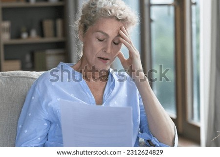 Shocked senior elder woman holding paper legal document, sitting on home sofa, touching head with closed eyes, feeling stress, despair, thinking on bad health problems, bankruptcy, financial crisis