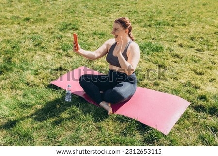 Image of young fitness woman sitting in gym while using phone. Beautiful sports blogger sitting on sport mat after practicing yoga on grass background