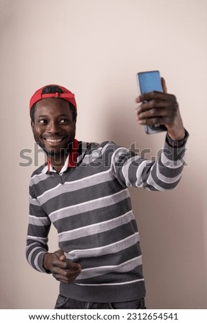 A young man with a trendy outfit and a cap is capturing selfies using his smartphone. The studio background provides a clean and neutral setting. Perfect for modern lifestyle, technology,