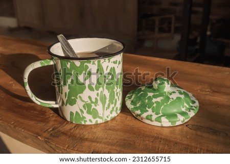 Hot tea or coffee in an enameled iron glass with a green abstract pattern. Old household appliances in wooden table with sunlight. Concept for retro, vintage, old fashioned, classic, art.