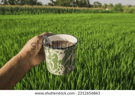 Someone is relaxing and holding hot tea or coffee in an enameled iron glass with a green abstract pattern and meadow rice field background. Concept for retro, vintage, old fashioned, classic, art.