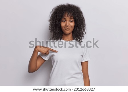 Look at me. Pleased dark skinned woman points at copy space on her blank t shirt for your promotional content brags about herself isolated over white wall boasts of her success. Place your design here Royalty-Free Stock Photo #2312668327