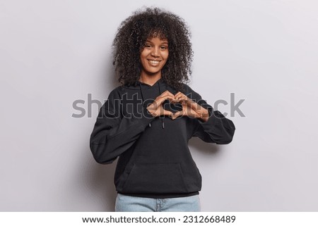 Horizontal shot of curly haired African girl makes heart gesture demonstrates her true feelings being in love with someone smiles gladfully wears casual black hoodie isolated over white wall