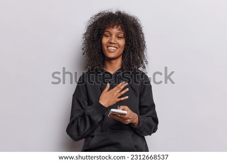 Positive curly haired teenage girl keeps hand on chest smiles pleasantly feels thankful holds mobile phone glad to receieve messages from boyfriend isolated over white background. Thank you very much Royalty-Free Stock Photo #2312668537