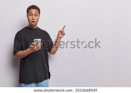 Studio shot of shocked speechless man stares with omg expression at camera points index finger at blank space holds mobile phone dressed in casual black t shirt and jeans isolated on white background Royalty-Free Stock Photo #2312668549