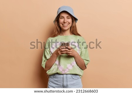 Indoor shot of good looking woman uses cell phone messages being happy to text with friends wears panama t shirt and jeans isolated over brown background. People and modern technologies concept