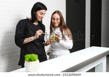 Medical staff. Young female doctor aesthetician with medical assistant using smartphone, checking list of appointments, standing behind a reception desk in modern white interior of a medical clinic Royalty-Free Stock Photo #2312690469
