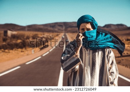 Portrait of a young white man dress as berber in the desert, blue scarf cover his face and a long grey and white cloth cover his body, photo in Sahara