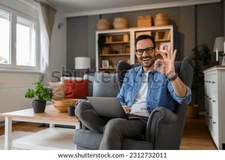 Portrait of cheerful male freelancer with laptop sitting comfortably on armchair and showing OK sign. Happy young entrepreneur gesturing and working over wireless computer remotely from home office