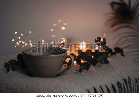 A cup with tea on a woolen blanket, a branch of eucalyptus and lights behind.