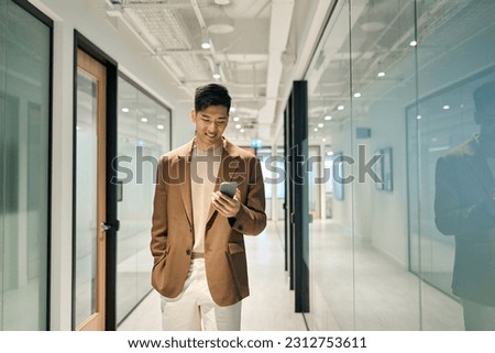 Young happy Asian business man using mobile cell phone tech standing in office hallway. Professional Japanese businessman holding smartphone, working on cellphone corporate device walking in office. Royalty-Free Stock Photo #2312753611