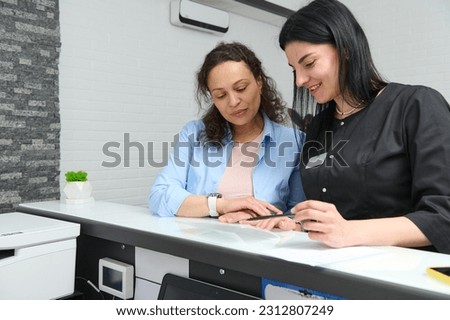Latin American beautiful woman, female patient at doctor appointment, discussing treatment, getting medical prescription, filling out and signing forms, standing by reception counter in modern clinic