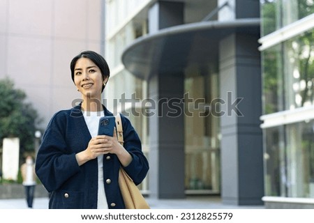 An Asian woman is using a smartphone app while walking on the street in front of an urban office building.