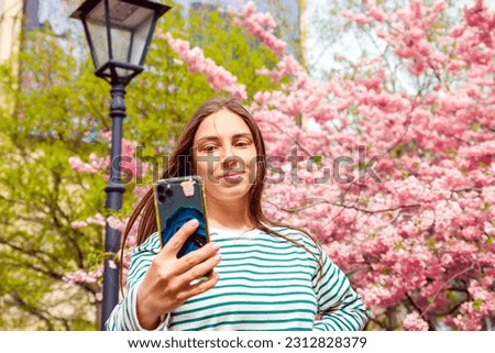 woman making video call, taking selfie portrait with mobile phone. Young smiling european white female woman on on cherry blossom flower sakura background, outdoor park.