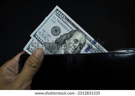 Someone hand holding two 100 one hundred dollars banknote bills inside black long wallet isolated on black dark background. Foreign currency, USD IDR, Money changer, trade, buy sell.