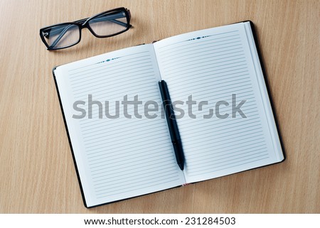 Open diary or office journal with a double page lined blank spread for your text with a ballpoint pen and glasses on a wooden desk, overhead view