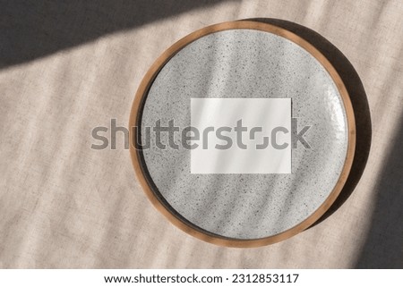 Minimalist business brand template, blank paper card mockup on ceramic plate, on a beige linen tablecloth background in sun light, wedding invitation or greeting card design