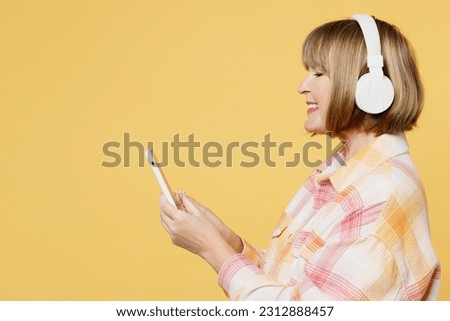 Side view elderly blonde caucasian woman 50s years old she wear casual clothes headphones listen to music use mobile cell phone isolated on plain yellow background studio portrait. Lifestyle concept