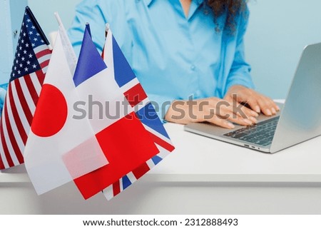 Cropped close up young successful employee business woman in casual shirt sit work at white office desk with pc laptop near many flags isolated on plain pastel light blue background studio portrait