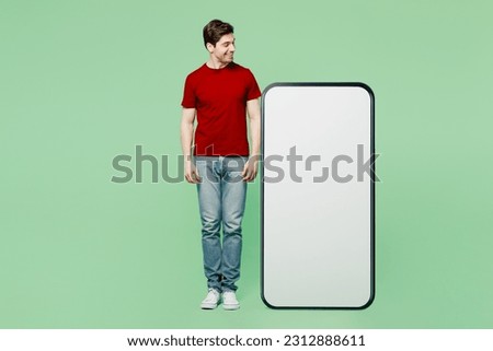 Full body happy smiling young man he wears red t-shirt casual clothes looking at big huge blank screen mobile cell phone smartphone with workspace area isolated on plain pastel light green background