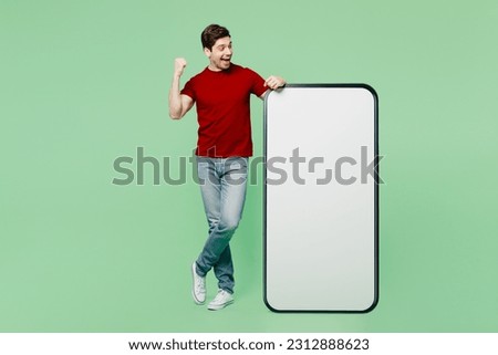 Full body happy young man he wears red t-shirt casual clothes big huge blank screen mobile cell phone smartphone with workspace area do winner gesture isolated on plain pastel light green background