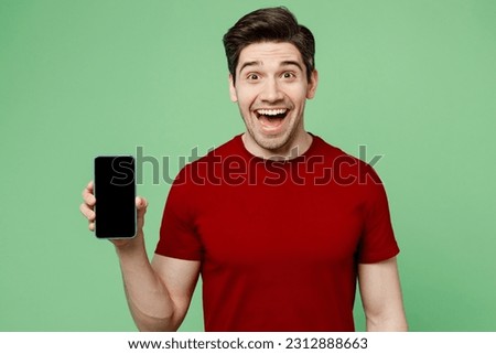 Young surprised shocked man he wearing red t-shirt casual clothes hold in hand use mobile cell phone with blank screen workspace area isolated on plain pastel light green background studio portrait