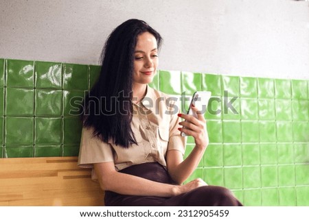 Photo portrait of girl holding phone and sitting on wooden bench in restaurant on green tile background. summer and spring concept, work remotely
