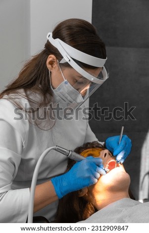 Female dentist and assistant removing dental calculus from teeth. Visit is in proffessional dental clinic. Woman sits on dental chair. Drilling and treatment of tooth, filling. Vertical photo Royalty-Free Stock Photo #2312909847