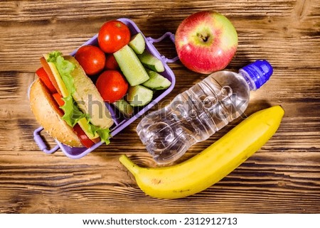 Ripe apple, banana, bottle of water and lunch box with hamburger, cucumbers and tomatoes on rustic wooden table. Top view