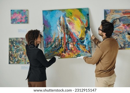 Couple of employees hanging paintings on the wall, they preparing for art exhibition in gallery