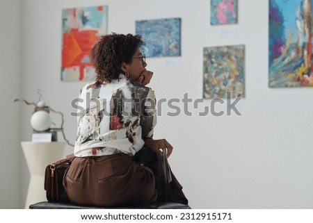 Rear view of African American woman sitting on couch and looking at paintings on the wall in gallery Royalty-Free Stock Photo #2312915171