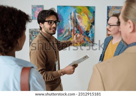 Young guide pointing at paintings on the wall and talking about modern art to people in gallery
