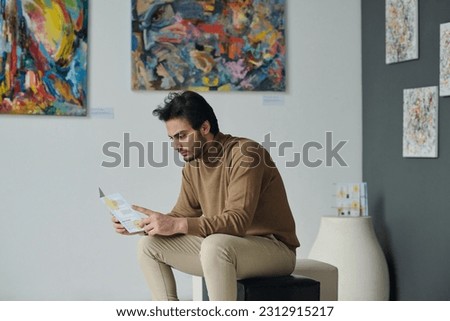 Young man resting on couch and reading brochure at exhibition of modern art in gallery