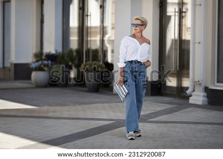 Rejuvenated Trendy mature woman with short hair stands outdoors in city street wears youth clothing white shirt, jeans cargo pants, clutch. Urban style fashion, age and timeless beauty. Royalty-Free Stock Photo #2312920087